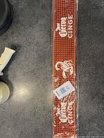 Long orange rubber bar mat with white 'Cinzano' and 'Cinge' text and scorpion graphic, sealed in original plastic packaging.
