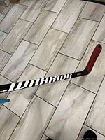 Full view of the left-handed carbon fibre hockey stick with visible 'WARRIOR' brand text and red taped blade on tiled floor