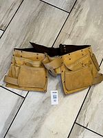 Full front view of the tan leather tool belt laid flat, showing multiple pockets and an adjustable brown strap.