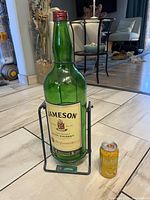 Large green glass Jameson Whiskey bottle standing in a black metal cradle stand on tile floor, next to a small soft drink can for scale.