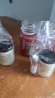 Photo showing four candles on a wooden surface including one red Yankee Candle brand jar candle and three cream-colored candles with black lids, two wrapped in plastic with a small matchstick jar each.