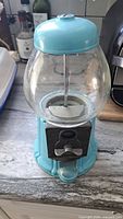 Front view of the vintage blue carousel style gumball machine showing metal base and top with glass globe, coin slot, and dispenser handle.