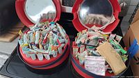 Two open red circular carrying cases holding many small paint tubes in assorted colors arranged inside, placed on a black surface.