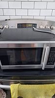 Hamilton Beach microwave oven on a stove with a stainless steel finish and digital display showing clock and timer. Microwave door with horizontal handle and a keypad control panel on the right side.