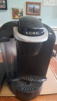 Front view of black Keurig coffee maker showing the machine's lid, branding, drip tray and water reservoir.