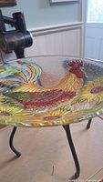 Close-up showing colorful textured glass basin with rooster and sunflowers, and black metal stand leg.