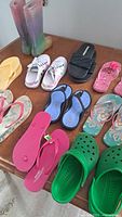Overview of women's footwear in various styles and colors, arranged on a wooden surface. Includes rain boots, flip-flops, clogs, slides, and sneakers.
