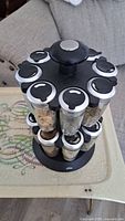 View of the rotating spice rack with 16 glass spice jars with black sprinkle top lids. The containers hold various dried spices and herbs.