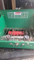 Front view of vintage Coleman camp stove showing two burners, red fuel tank, and metal grate. Green lid open revealing Coleman branding and usage instructions.