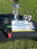 Full lot displayed on outdoor table showing Jack Daniels gift set in metal box with two shot glasses, plastic bar spouts, metal Man Cave sign, two beer steins, and thermometer.
