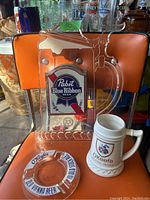 Photo showing Pabst Blue Ribbon beer sign, O'Keefe beer stein, and O'Keefe Old Vienna Beer ashtray on orange chair.