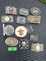 Photo of 12 assorted belt buckles on a black woven background showing logos and decorative styles.