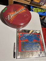 Full view of round Budweiser stool seat and rectangular Budweiser mirrored sign on white table with Sharpie pen for scale.