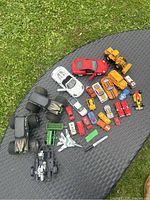 Wide view of all 22 metal toy cars, trucks, monster trucks, motorcycle trailer, and camouflage jet airplane spread on a textured outdoor table surface.