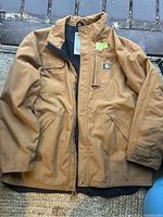 Front view of brown Carhartt jacket laid flat, showing zipper, pockets, and logo patch.