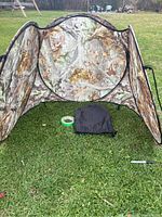 Pop up duck blind set up on grass showing circular opening, side wings, black carry case, and green tape.