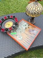 Photo showing the roulette wheel, chess/checkers glass case, and decorative lamp all placed on a black woven table outdoors.