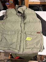 Roots puffer vest front view showing two large pockets with flap and light olive/khaki color, some dirt marks visible