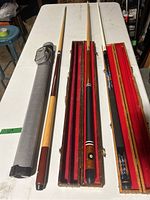 Three pool cues laid out next to their cases, two wooden and one soft gray case.