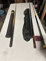 Photo showing Viking 8/9 ball cue, two cases, and a wooden green wrapped cue all laid on a white table.