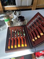Two sets of vintage stainless steel cutlery with faux wood handles displayed in original cases with additional glassware and kitchen items in background.