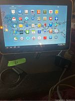 Samsung Galaxy Tab powered on showing home screen with multiple app icons, charger and cables visible in image.
