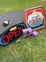 Overall view of the lot on outdoor table and grass, showing OPEN sign, Molson Canadian bar sign, and Crown Royal boxed glasses.