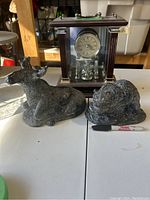 Two soapstone animal sculptures in front of a wooden mantle clock with glass panels on a white surface.