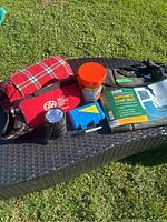 Full lot overview on wicker chair: camp seat packaging, red plaid blanket, red CAA cooler bag, orange OtterBox container, blue water safety kit case, and flashlight.