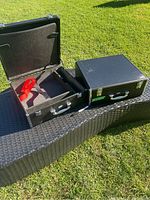 Two black plastic storage cases with foam padding and metal latches shown outdoors on a woven black surface. One case is open showing foam interior with red Velcro strap and compartments.