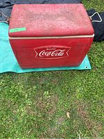 Full front view of the red vintage Coca Cola cooler on a green tarp showing the Coca Cola logo and weathering on the lid.
