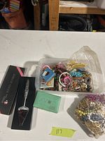 Plastic container with variety of vintage jewelry items including rings, bracelets, earrings, and an Avon item along with the boxed cheese slicer.