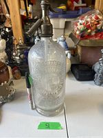 Full view of the vintage etched glass seltzer bottle showing cylindrical shape, thickness, and metal siphon on top with lever dispenser.