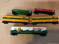 Five HO scale model train cars arranged on a surface: two green CP Rail gondola cars, two yellow and green ATSF boxcars, one green and white Star-Kist flatcar with trailer.