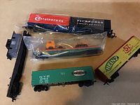Six HO scale vintage train cars displayed on a beige surface, including a black and red container car, a green NYC boxcar, a yellow Heinz freight car, a black caboose, and a green gondola car inside plastic packaging with miniature vehicles.