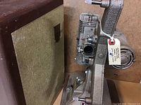 Side view of the Keystone 8mm film projector K109 showing the film gate and motor housing attached to the base, with carry case behind.