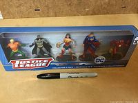 Front view of sealed Justice League 5-pack box containing Aquaman, Batman, Wonder Woman, Superman, and Flash figurines, with a Sharpie for scale.