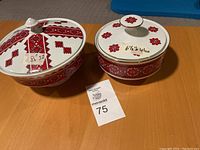 Two porcelain serving dishes with lids side by side on wooden table, one larger (8.5 in) one smaller (6.5 in wide). Both decorated with red folk patterns and gold trim.