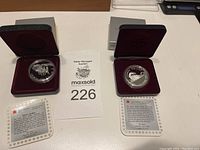 Two silver coins displayed in red velvet-lined cases with certification cards visible for each coin.