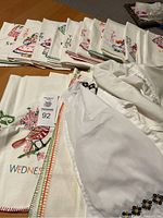 Stack of 27 embroidered dishtowels including day of the week designs visible, plus two white embroidered aprons on top