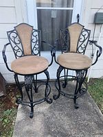 Pair of black iron and beige upholstered swivel barstools shown from the front on concrete and grass surface.
