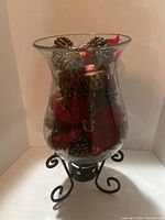 Full view of large clear glass vase sitting on black metal stand with Christmas decorations inside including pinecones, red poinsettia leaves, and red and gold ornaments.