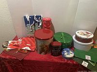Various sizes of Christmas-themed tin boxes and gift bags arranged on a red cloth, showing condition and designs.