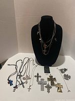 Mixed group of cross and angel pendant necklaces arranged on black necklace display stand and flat surface. Shows variety of styles and metals including black, silver-tone, gold-tone, and pewter finishes.