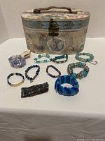 Jewelry box with butterfly artwork showing multiple compartments and clasp, surrounded by nine stretch bracelets of varied style and colors including Guess and Mogo brand bracelets.