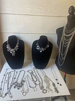 Full view of multiple silver tone necklaces displayed on black busts and mannequin including large beads, linked rings, and layered chains with various pendant styles