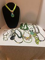 Photo showing 12 assorted green necklaces displayed on a white surface with one necklace on a black fabric display stand. Various materials include fabric, beads, metal, and shell. Condition shows wear and scratches.