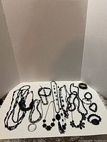 13 assorted black costume jewelry pieces including necklaces, chokers, and bracelets arranged on white backdrop. Variations in bead size, style and pendants visible.