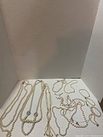 Top down view showing 11 assorted pearl and crystal necklaces and 4 pearl bracelets arranged side by side on white surface.