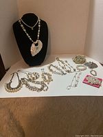 Overall photo of the entire collection of 14 costume jewelry pieces, including 9 necklaces and 5 bracelets laid flat and displayed on a black stand and white surface.